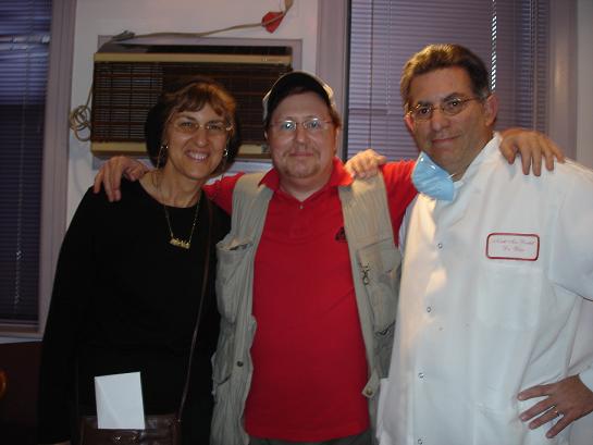 In the image, there are three individuals posing for a photograph two adults standing on either side of another adult who appears to be seated. The central figure is wearing a white lab coat and glasses, suggesting they might be a scientist or medical professional. The person in the middle is smiling and seems to be in a jovial mood, while the other two individuals are also smiling. They all appear to be in an indoor setting with a window visible in the background.